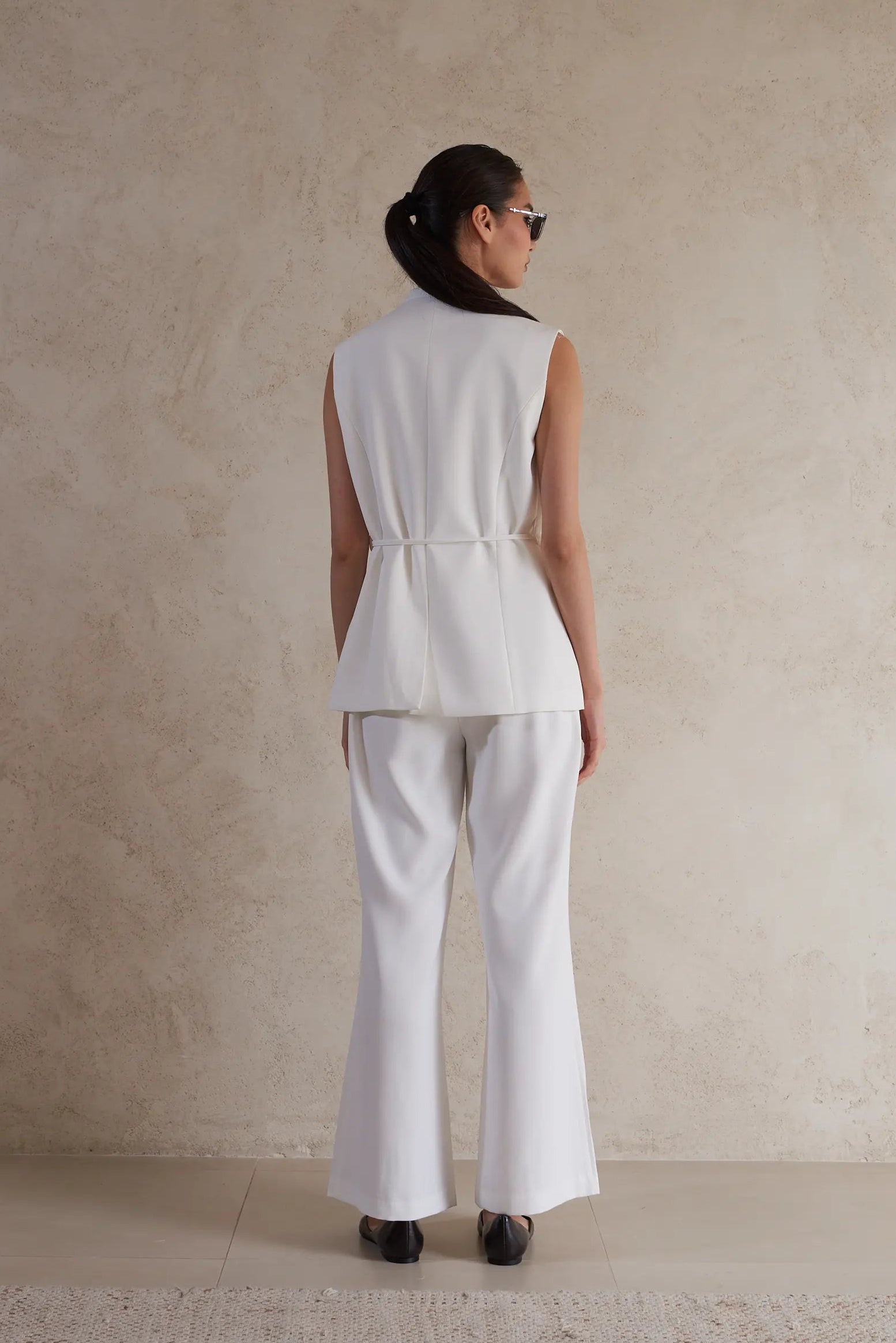 Person wearing a white sleeveless top and pants against a beige wall.